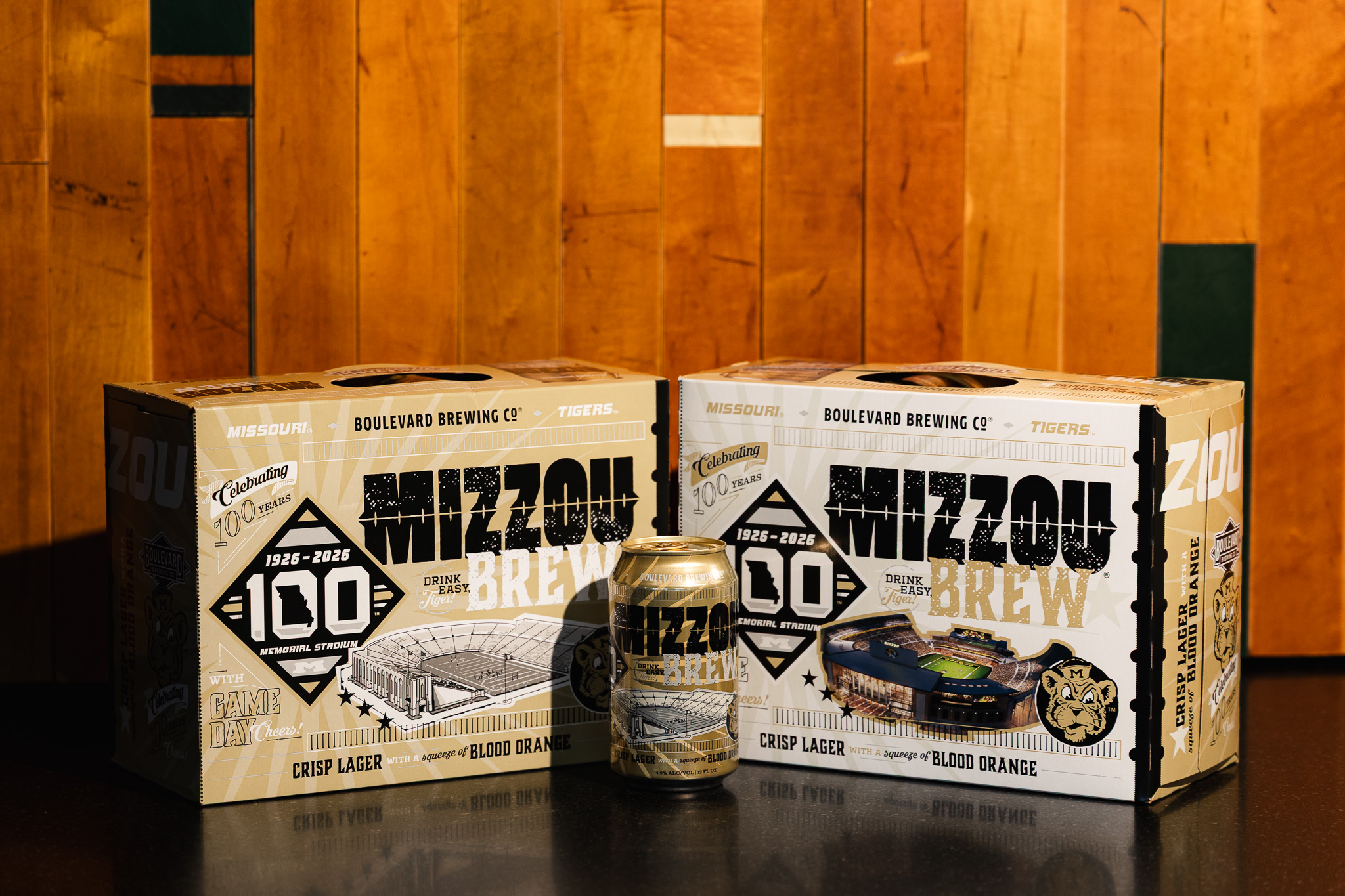 Two boxes and a can of Mizzou Brew beer are displayed on a wooden surface. The packaging features gold, black, and white colors with text “Mizzou 100”, celebrating Missouri’s centennial.