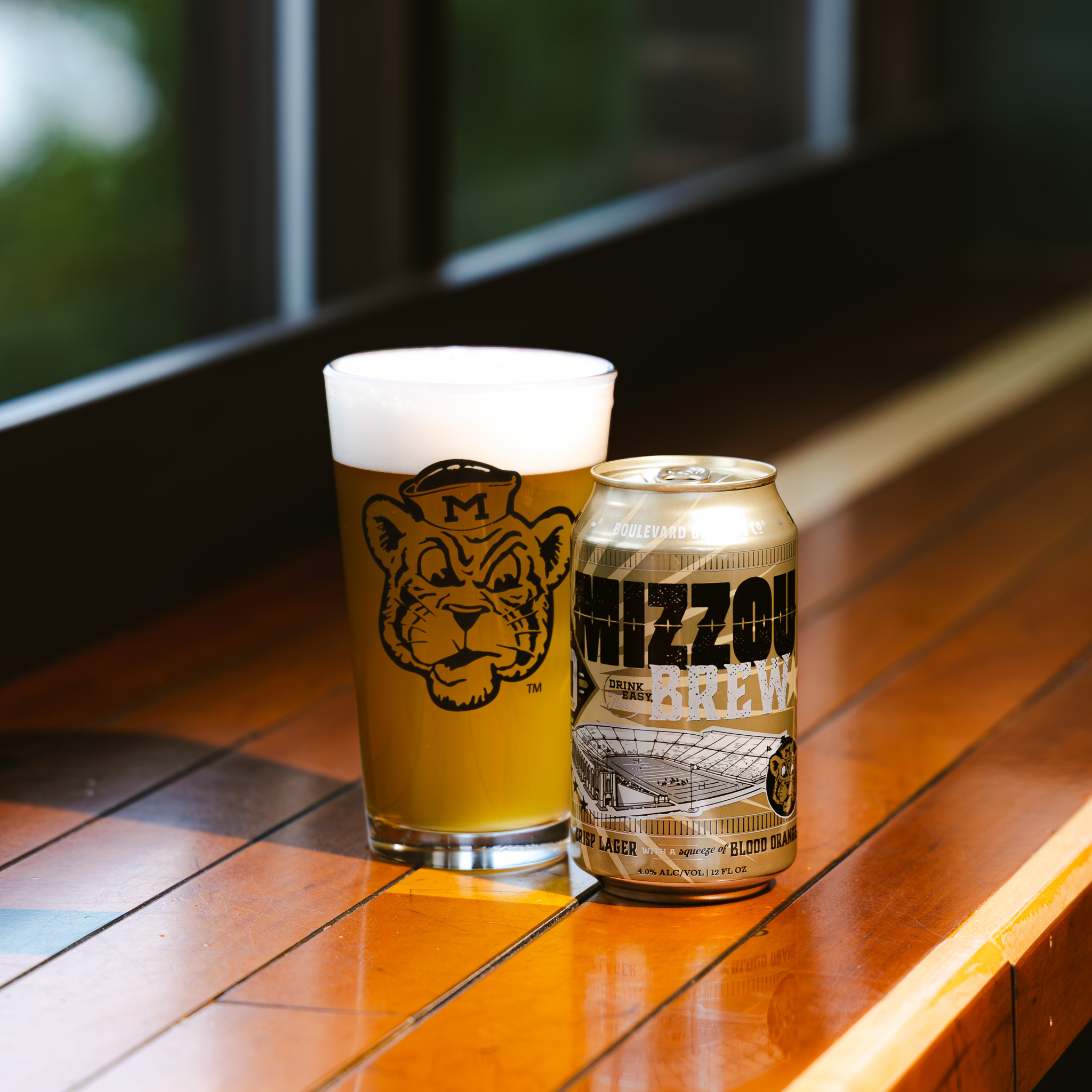 A can of Mizzou Brew and a pint glass filled with beer, both featuring a tiger mascot, sit on a wooden surface by a window with sunlight streaming in.