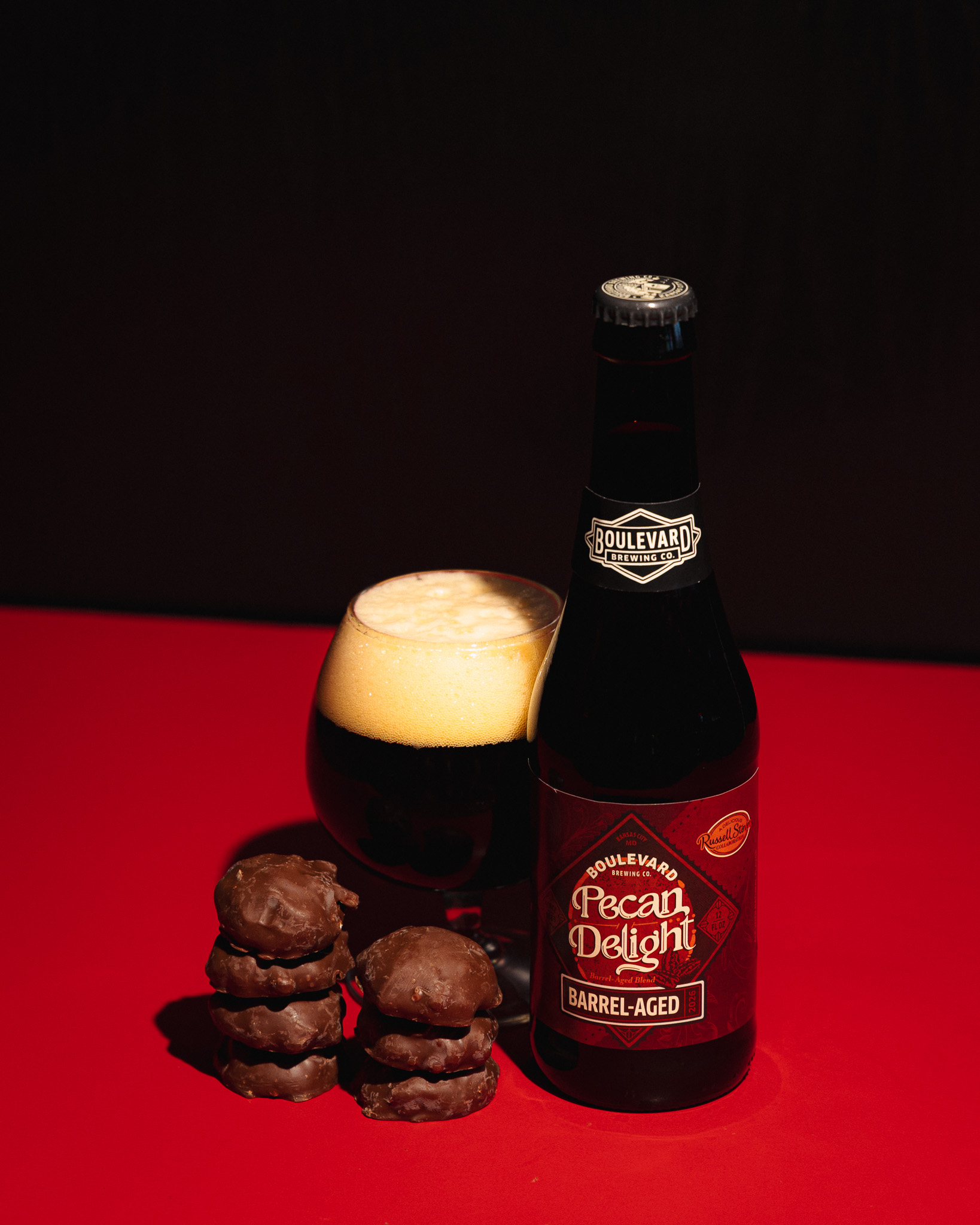 A bottle of Boulevard Pecan Delight Barrel-Aged beer stands next to a glass of dark beer with foam, and a stack of chocolate-covered cookies, all set against a red background.