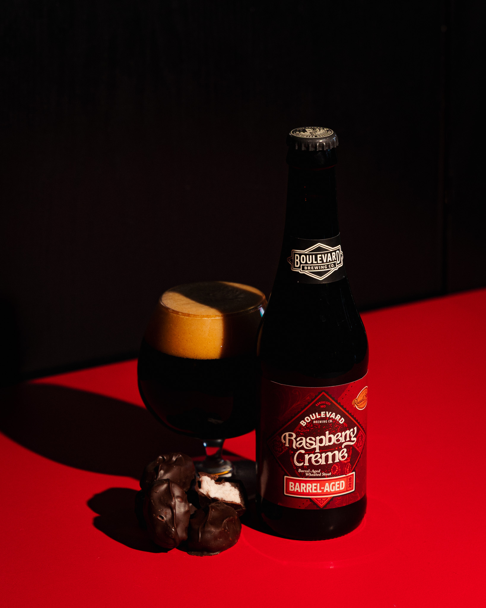 A bottle of Boulevard Raspberry Crème Barrel-Aged beer beside a filled glass and two chocolate-covered treats, all set on a bright red surface with a dark background.