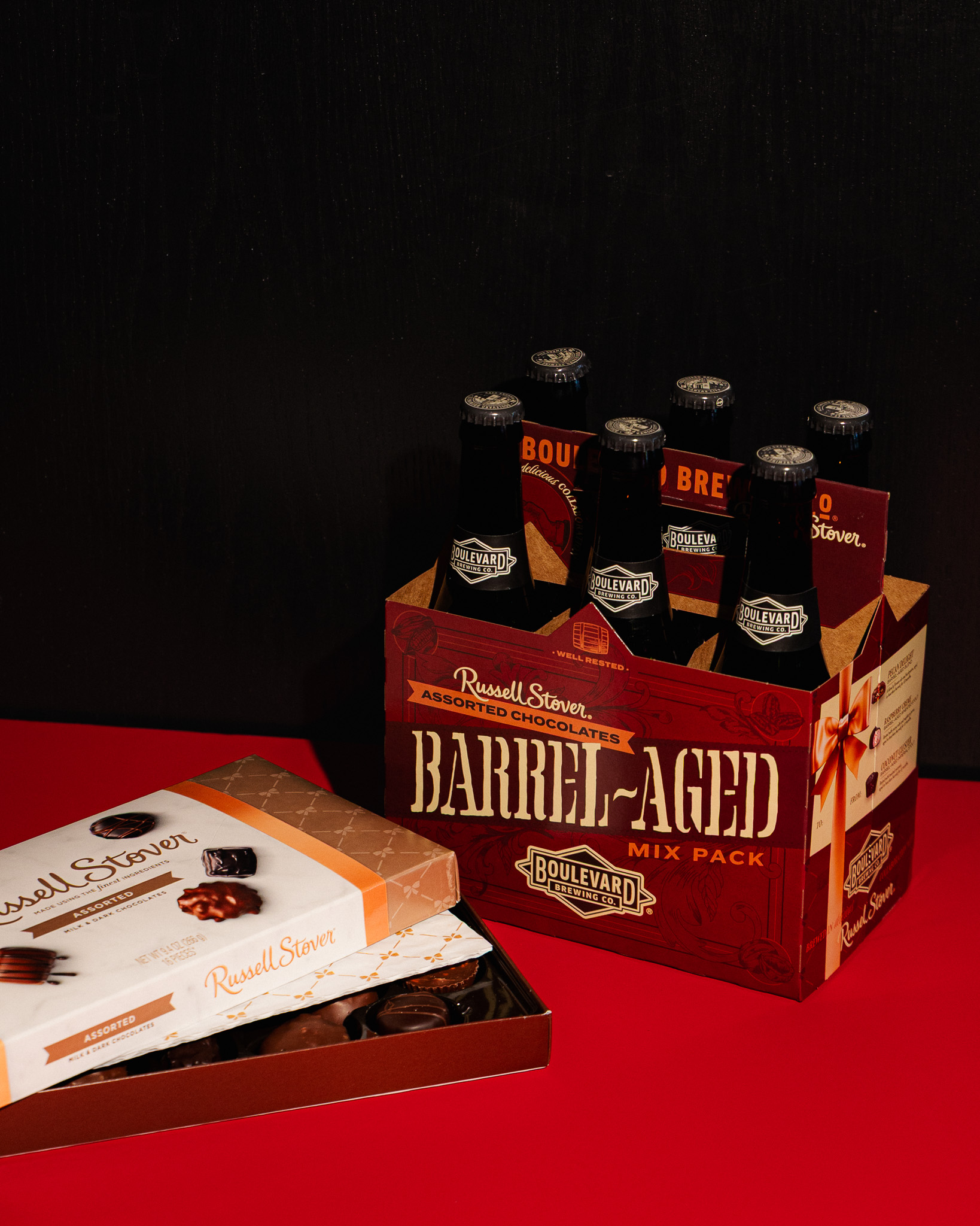 A six-pack of Boulevard beer with “Barrel-Aged” branding sits next to an open box of Russell Stover assorted chocolates on a red surface with a dark background.