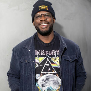 A smiling Black man with a beard, glasses, and a beanie that says "Grits & Eggs Podcast" wears a dark corduroy jacket over a black Pink Floyd graphic t-shirt, ready to bring his charm and wit to the stage of a comedy show, against a plain grey background.