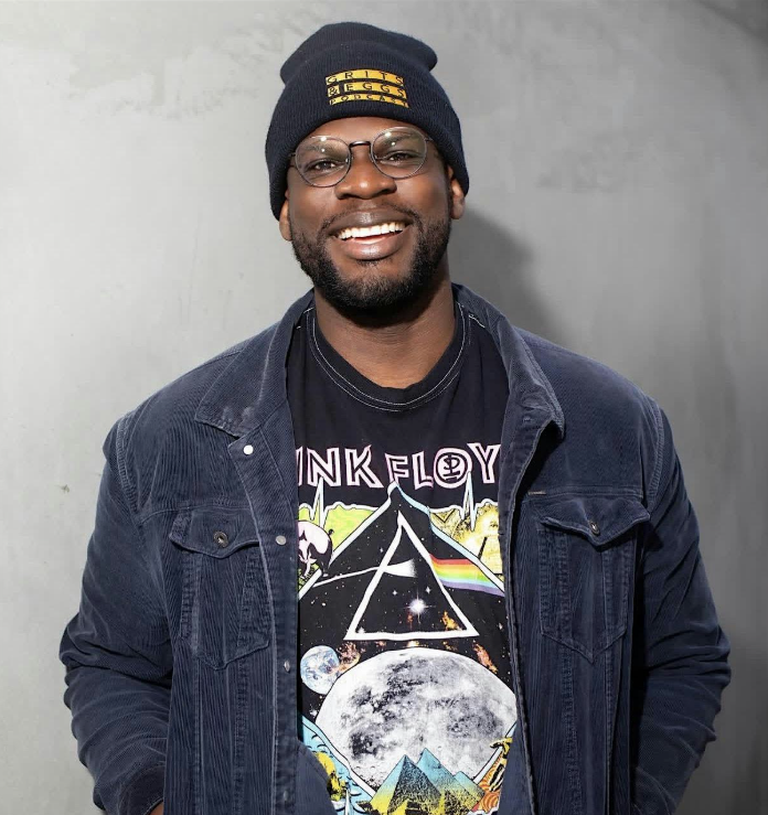 A smiling Black man with a beard, glasses, and a beanie that says "Grits & Eggs Podcast" wears a dark corduroy jacket over a black Pink Floyd graphic t-shirt, ready to bring his charm and wit to the stage of a comedy show, against a plain grey background.