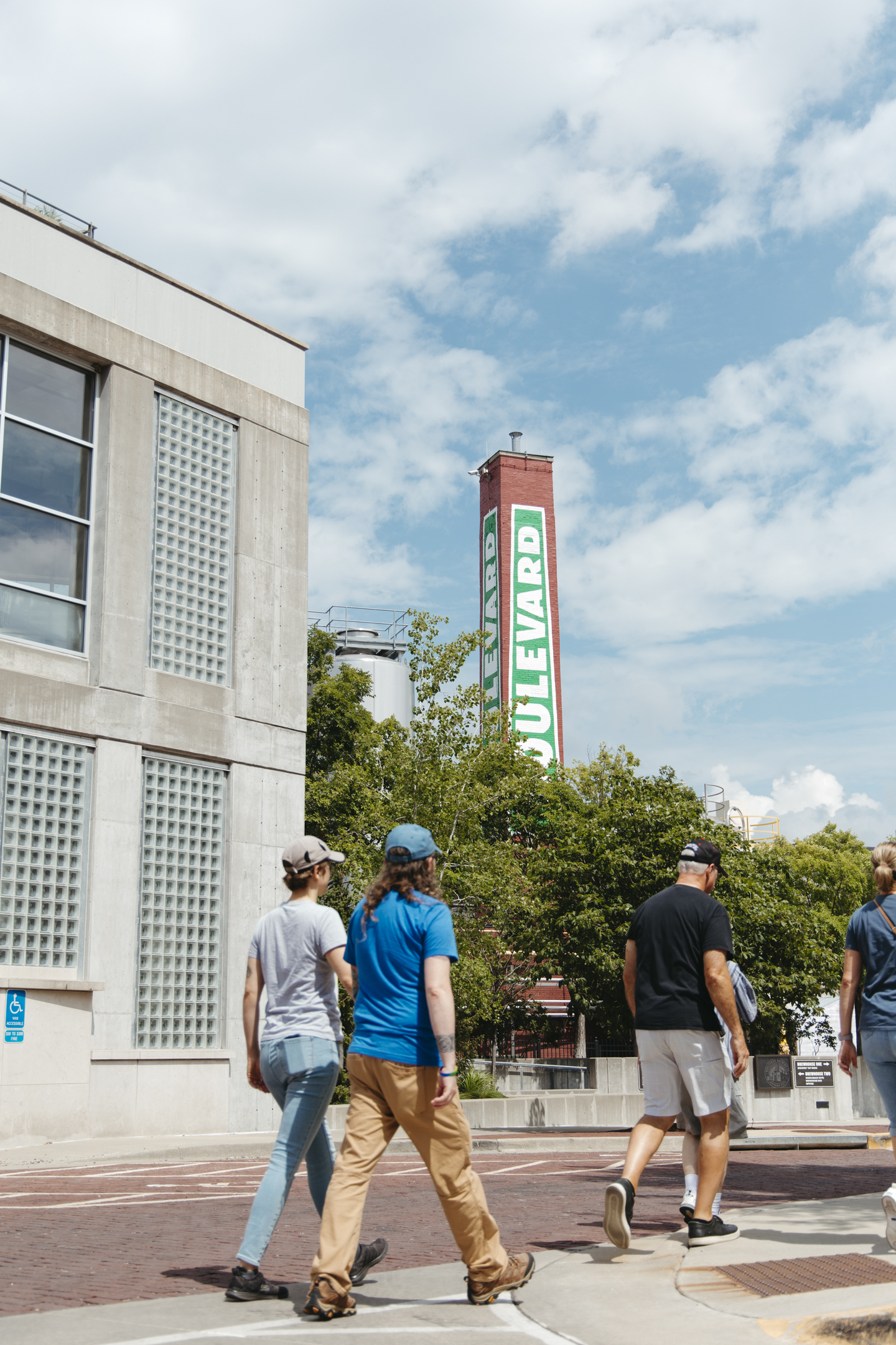 Four people cross a street near a tall sign reading "Boulevard" on a red structure, surrounded by modern buildings and green trees under a partly cloudy sky.