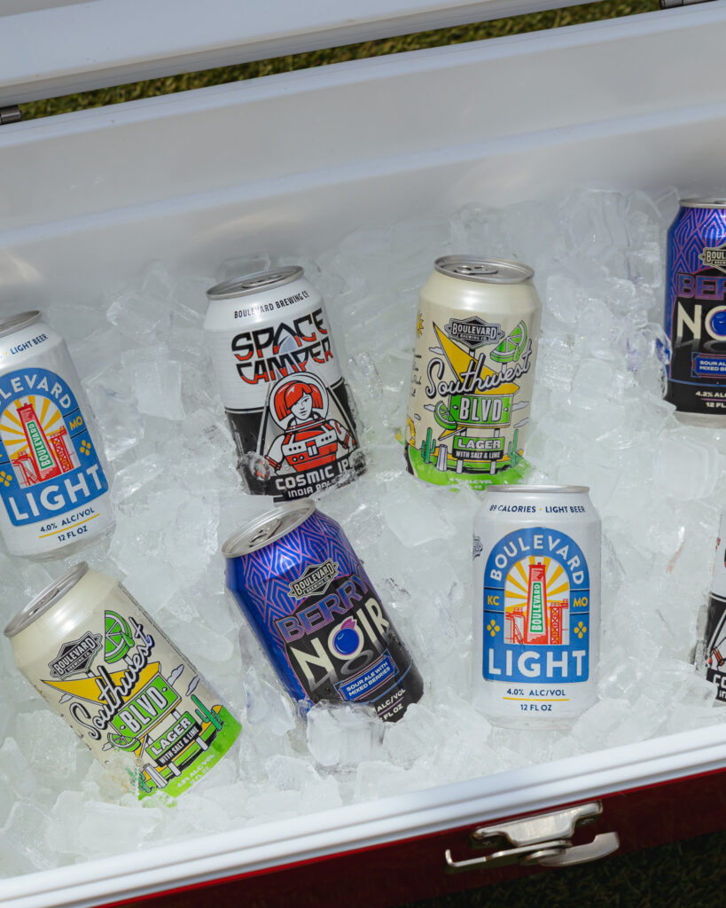 Cans of various beers from Boulevard Brewing Co. Kansas City, Missouri, sit upright on ice inside an open cooler, including popular varieties like Boulevard Light, Berry Noir, Space Camper, and Southwest Blvd. The cooler is filled with ice to keep the drinks refreshingly cold.