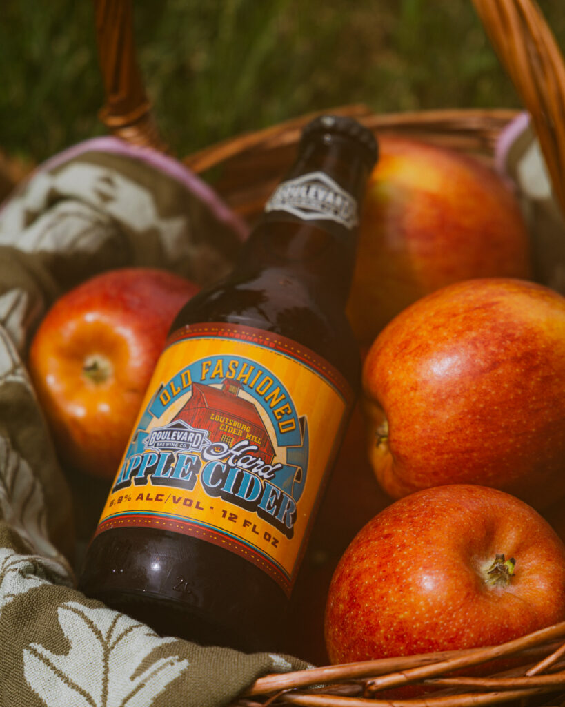 A bottle of Old Fashioned Hard Apple Cider rests in a wicker basket with three red apples and a patterned cloth, outdoors on grass—a perfect Boolevard-inspired arrangement for a crisp afternoon picnic.