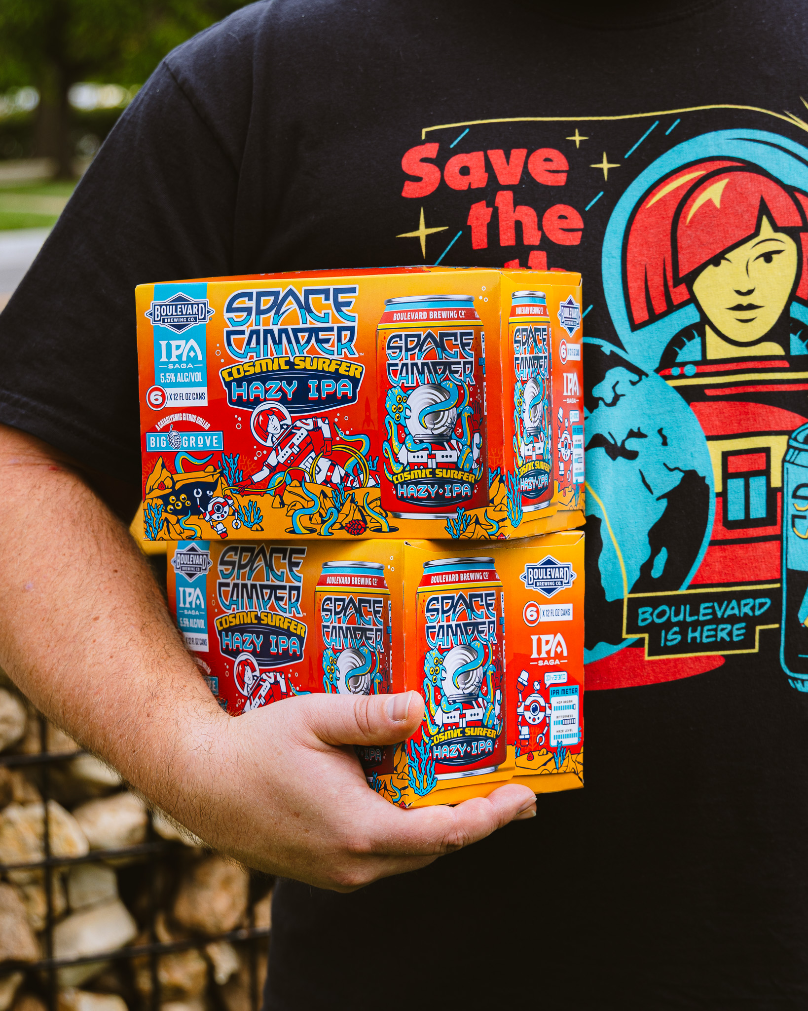 A person in a black “Save the Earth” t-shirt holds two six-packs of Space Camper Cosmic Surfer IPA beer outdoors. The colorful cans feature comic-inspired space artwork.