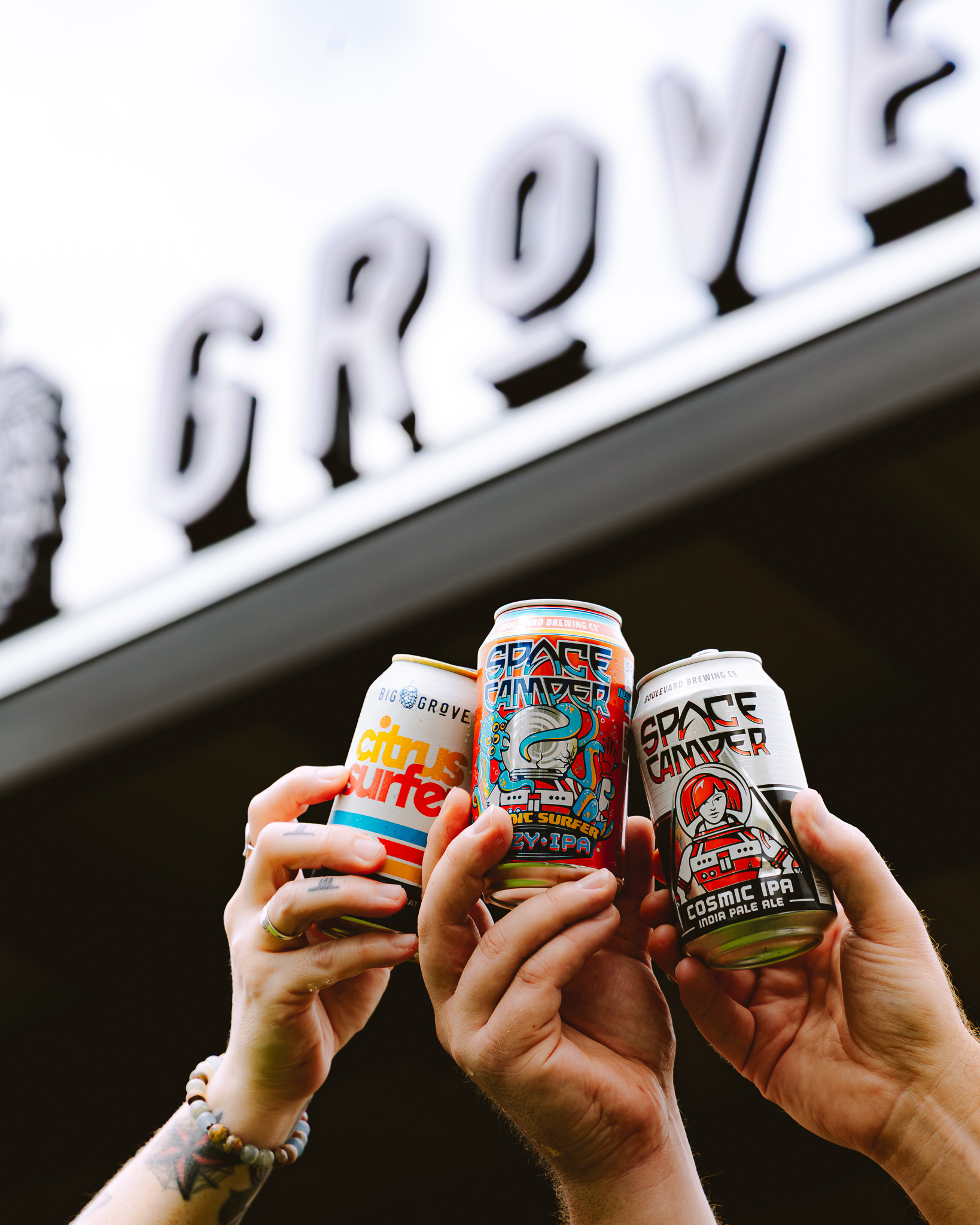 Three hands hold up colorful beer cans in front of a building with a partially visible sign that reads “GROVE.” The cans have unique designs and labels, each representing a different beer.