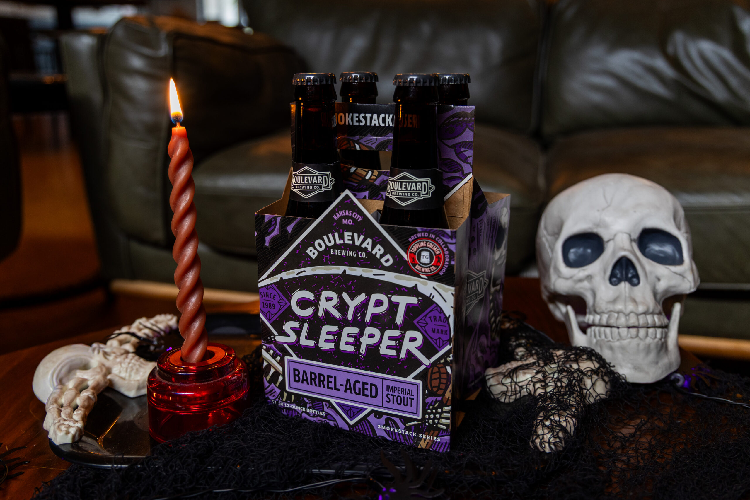 A six-pack of Boulevard Brewing Co. Crypt Sleeper Barrel-Aged Imperial Stout sits on a table with a lit twisted candle, a white skull, and Halloween-themed decorations.