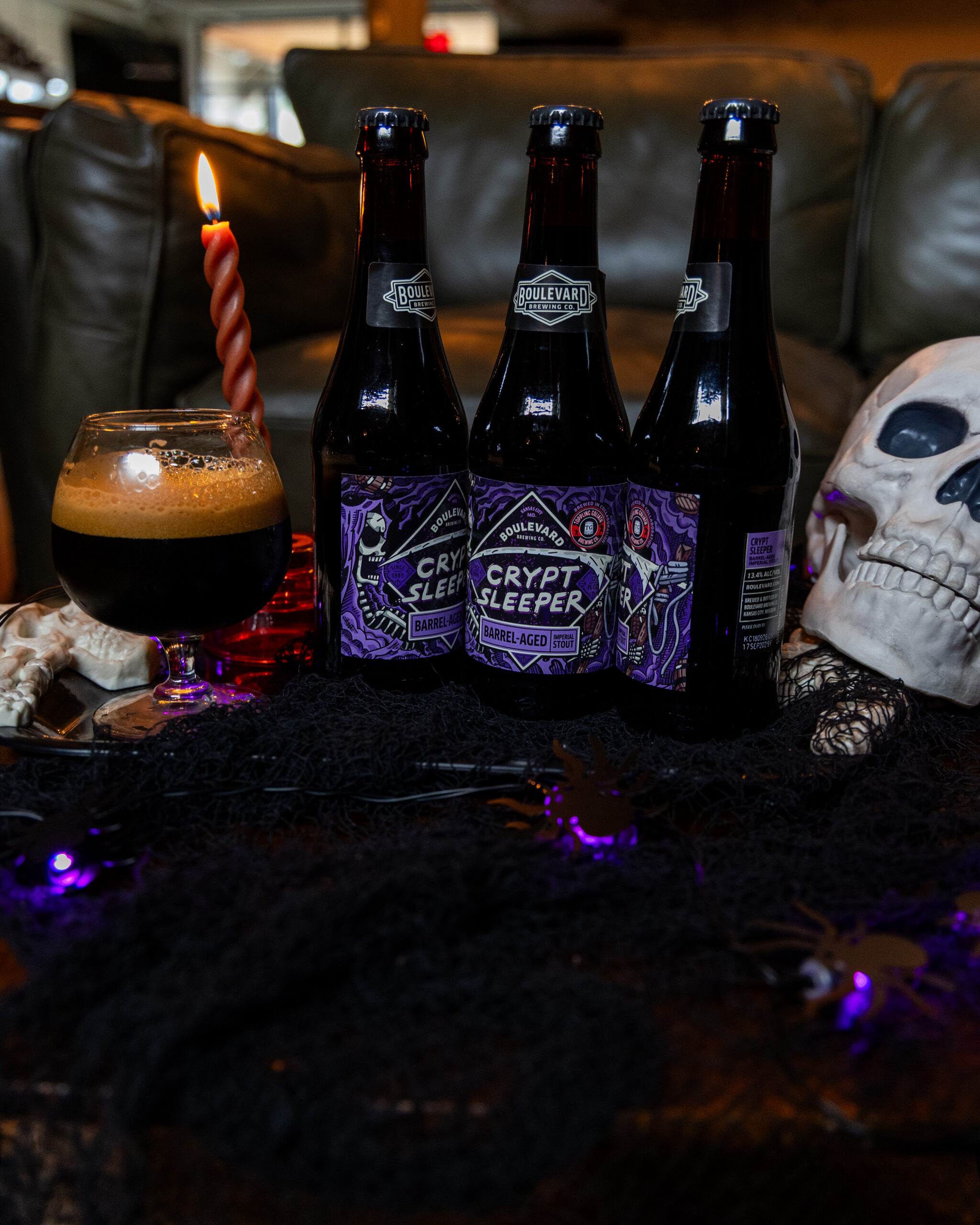 Three “Crypt Sleeper” beer bottles, a glass of dark beer, a twisted red candle, a skull, and Halloween decorations are arranged on a table with dark fabric and purple string lights.