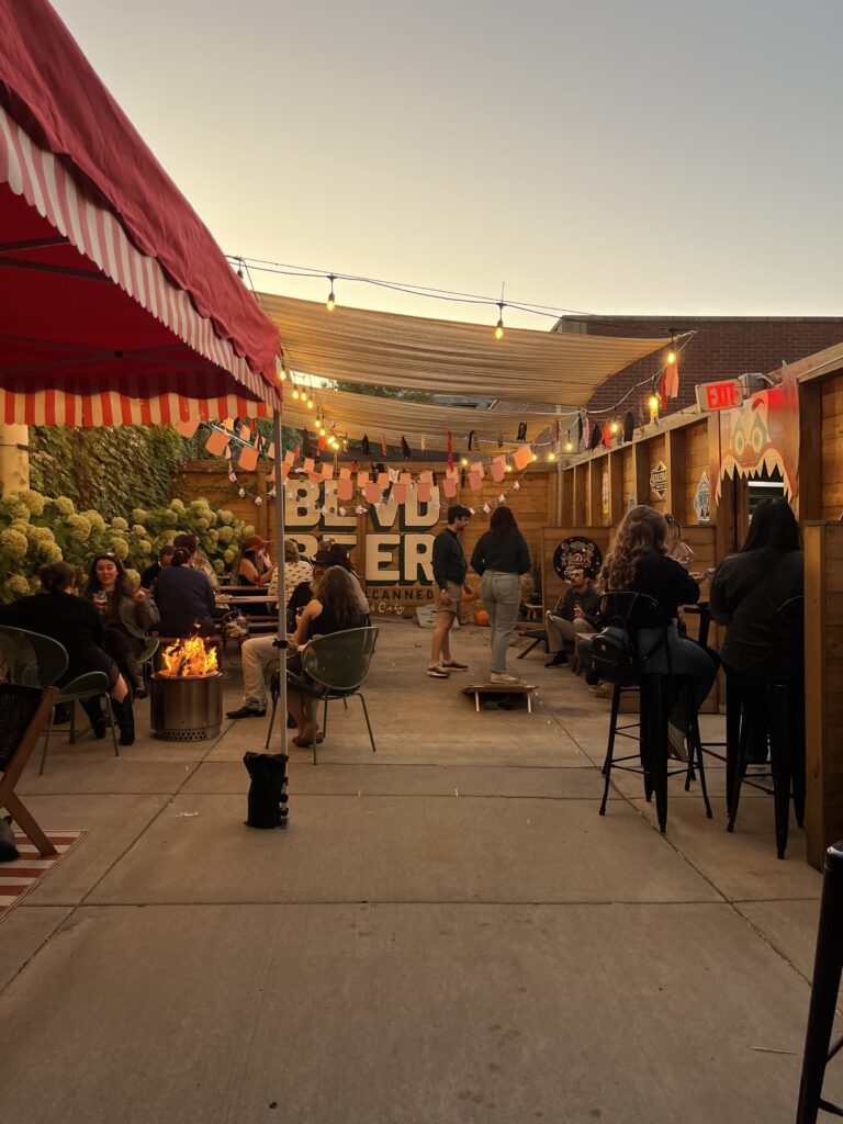 People sit and stand at an outdoor patio on the Boolevard, decorated with string lights and flags at sunset, some gathered around a fire pit. The atmosphere is casual and social.