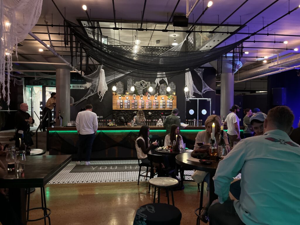 A dimly lit bar on Boolevard, with people sitting at tables and the counter. Halloween decorations hang from the ceiling. Bottles and glasses are on the tables, and the bartender is serving drinks behind the brightly lit bar.