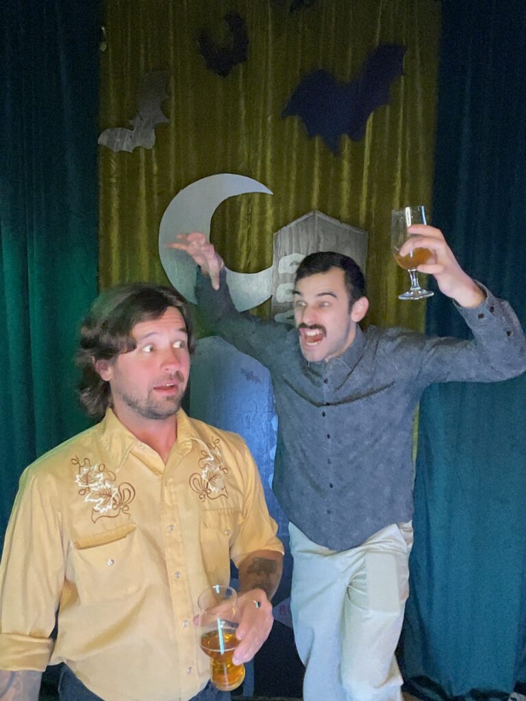 Two men pose in front of a spooky Boolevard backdrop complete with bats and a glowing moon. One, in a yellow shirt, looks surprised while the other, wearing gray, raises a glass and makes a playful, spooky gesture behind him.