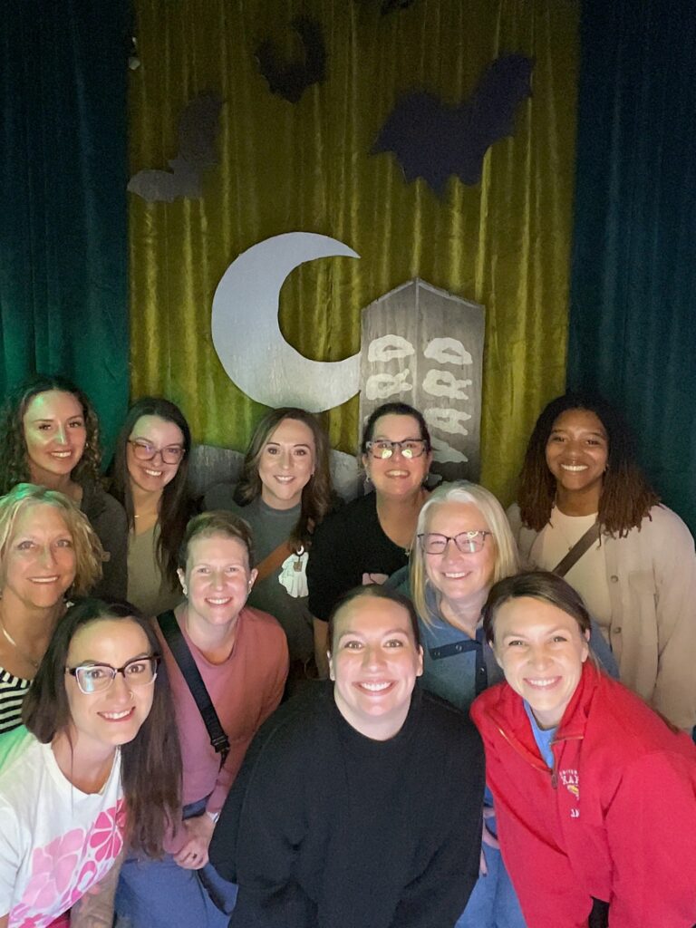 A group of eleven smiling women pose together indoors in front of a Boolevard-themed backdrop featuring a crescent moon, gravestones, and bats, suggesting a Halloween or spooky theme.