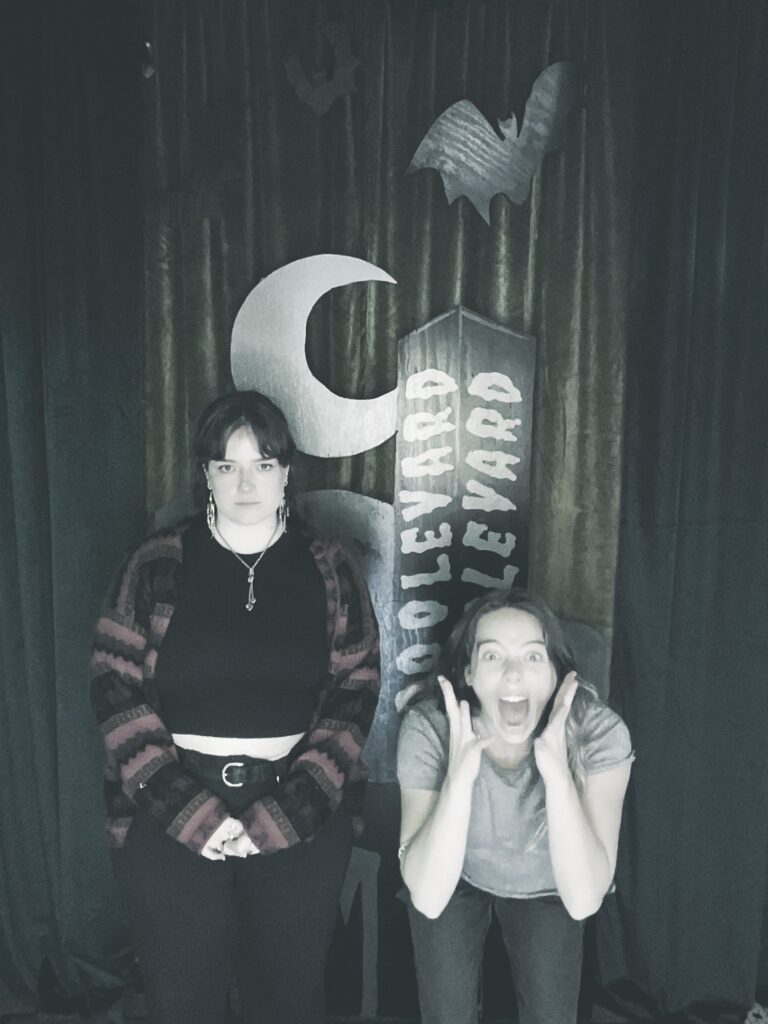 Two people pose in front of a spooky backdrop with a crescent moon, bats, and a tombstone reading “Boolevard.” One stands with a serious expression, while the other crouches with an excited, surprised face.
