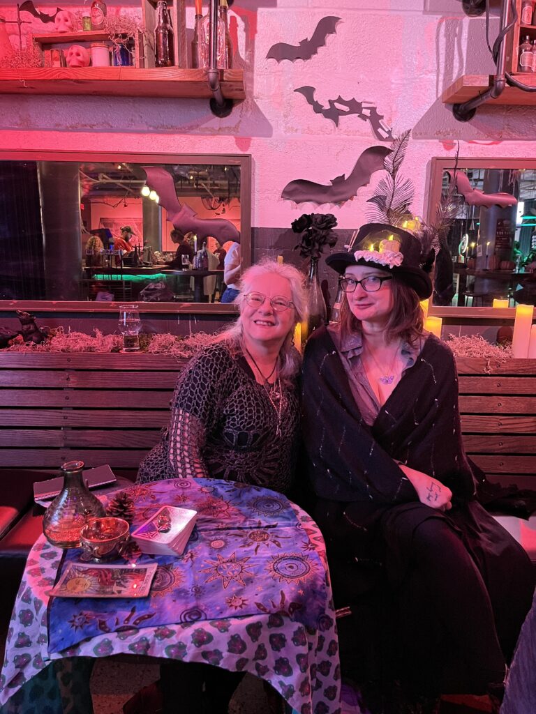 Two people sitting at a decorated table in Boolevard, a bar adorned with festive Halloween decor including bats on the wall. Both are smiling; one wears a black top hat with goggles and feathers, while the other sports glasses and patterned clothing.