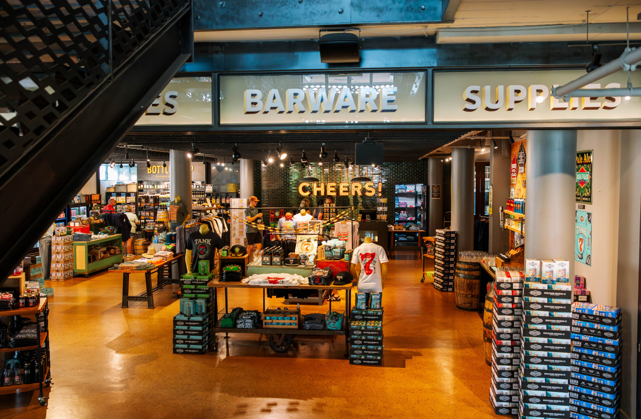 Interior view of the Boulevard Brewing Co. retail store featuring a “BARWARE” sign, a “CHEERS!” light-up display, and shelves stocked with apparel and stacked cases of beer.