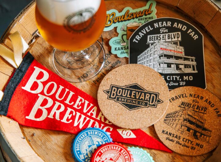 A glass of TBIF beer, a felt pennant, stickers, coasters, and bottle caps featuring the Boulevard Brewing Co. logo arranged on top of a wooden barrel.