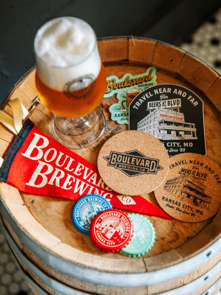 A glass of TBIF beer, a felt pennant, stickers, coasters, and bottle caps featuring the Boulevard Brewing Co. logo arranged on top of a wooden barrel.