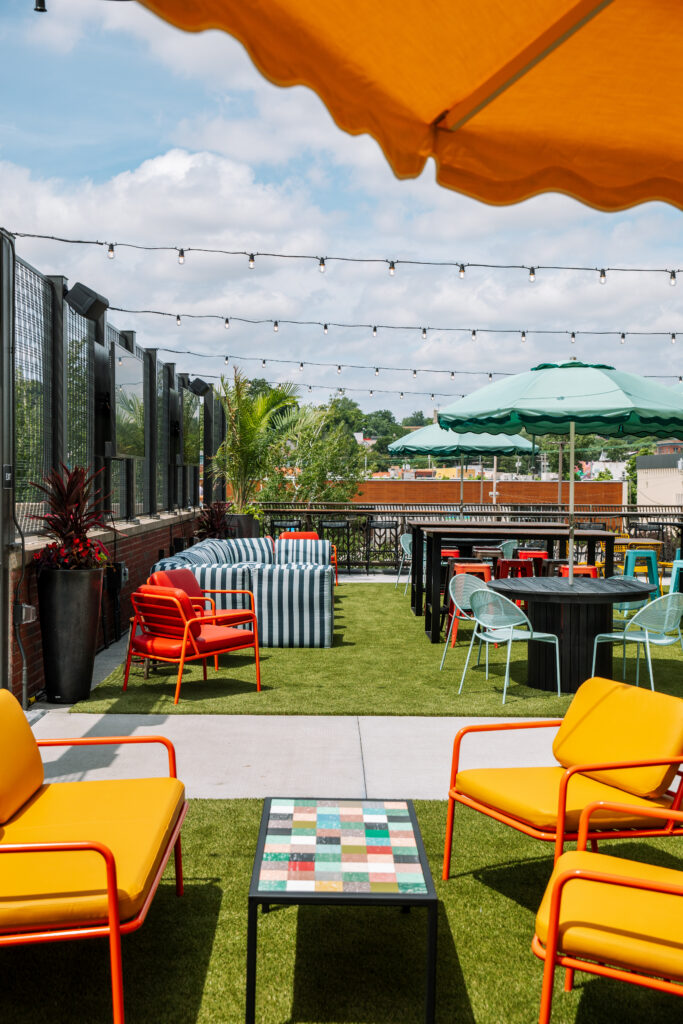 A rooftop terrace at Boulevard Brewing Co. in Kansas City featuring artificial turf, orange chairs, a striped sofa, dining tables, and colorful sun umbrellas under a string of lights.