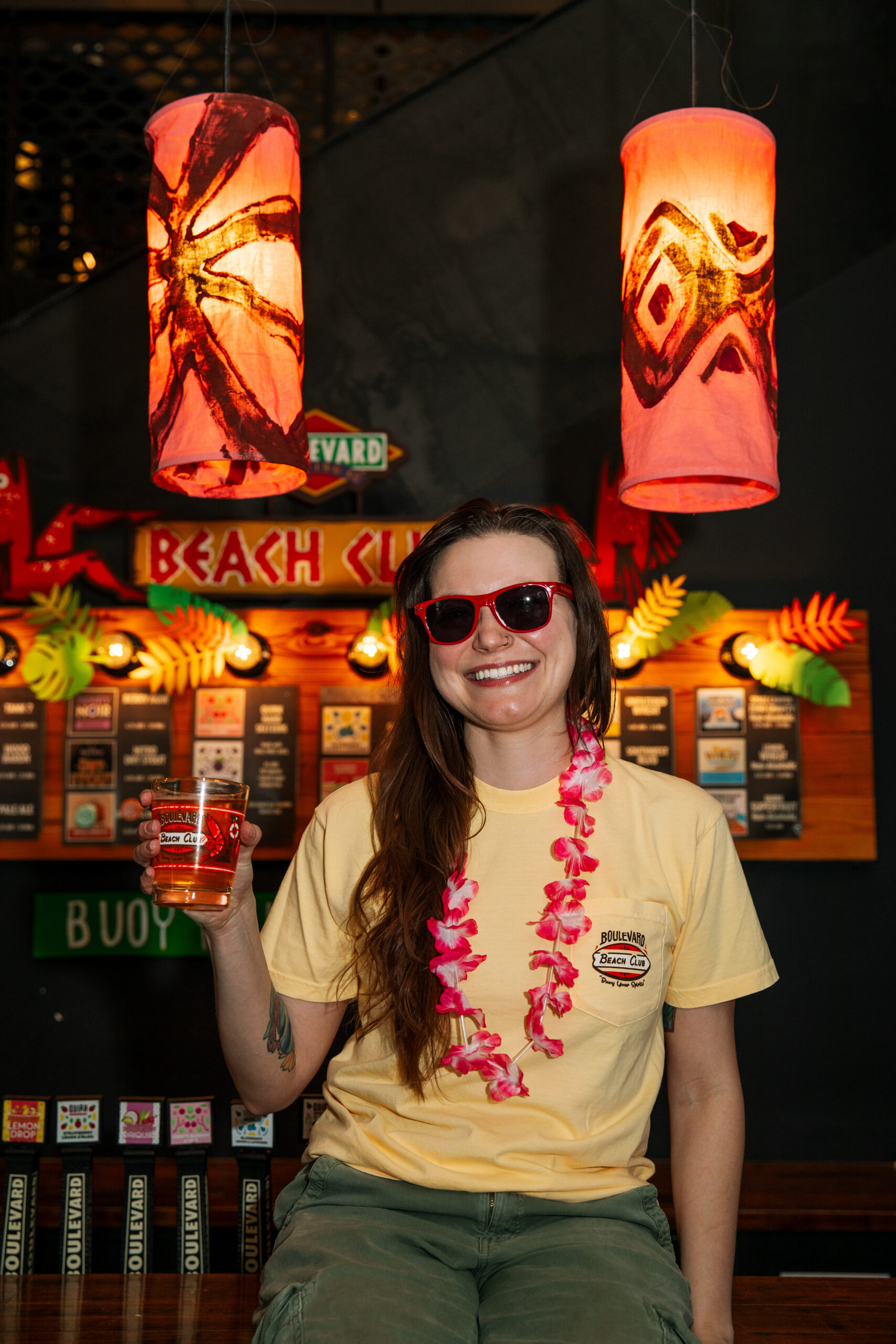 A woman wearing a yellow t-shirt, pink lei, and red sunglasses smiles while holding a glass of beer in front of a Beach Club bar backdrop.
