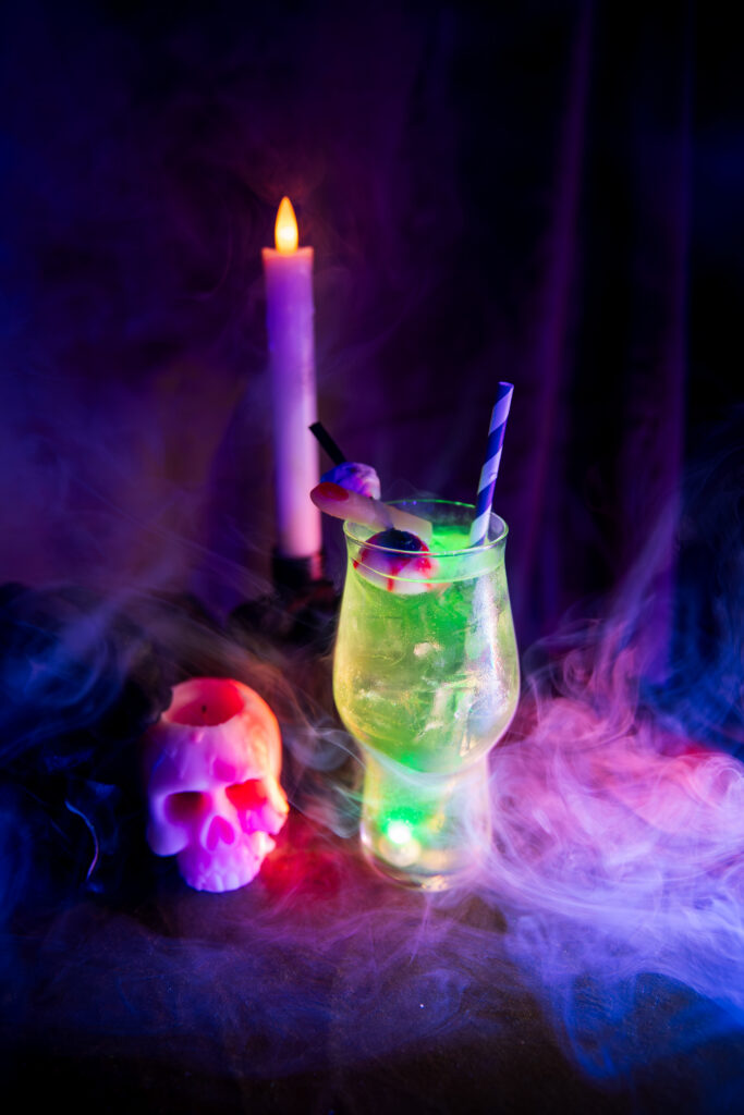 A Boolevard cocktail, green and garnished with an eyeball, sits among smoke, a skull candle, and a taper candle.