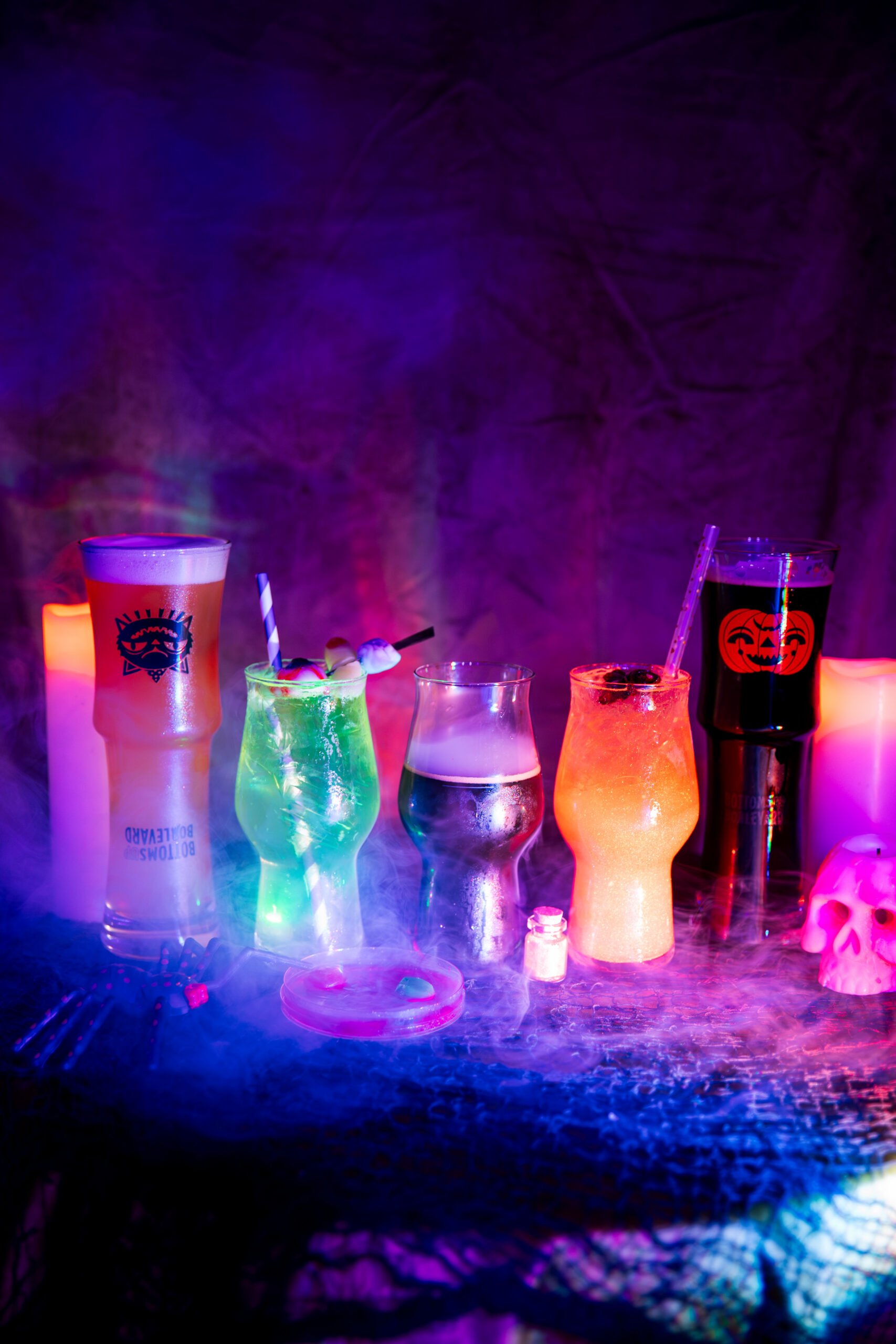 Five colorful Boolevard Halloween-themed cocktails in various glasses arranged on a table with purple and blue fog.
