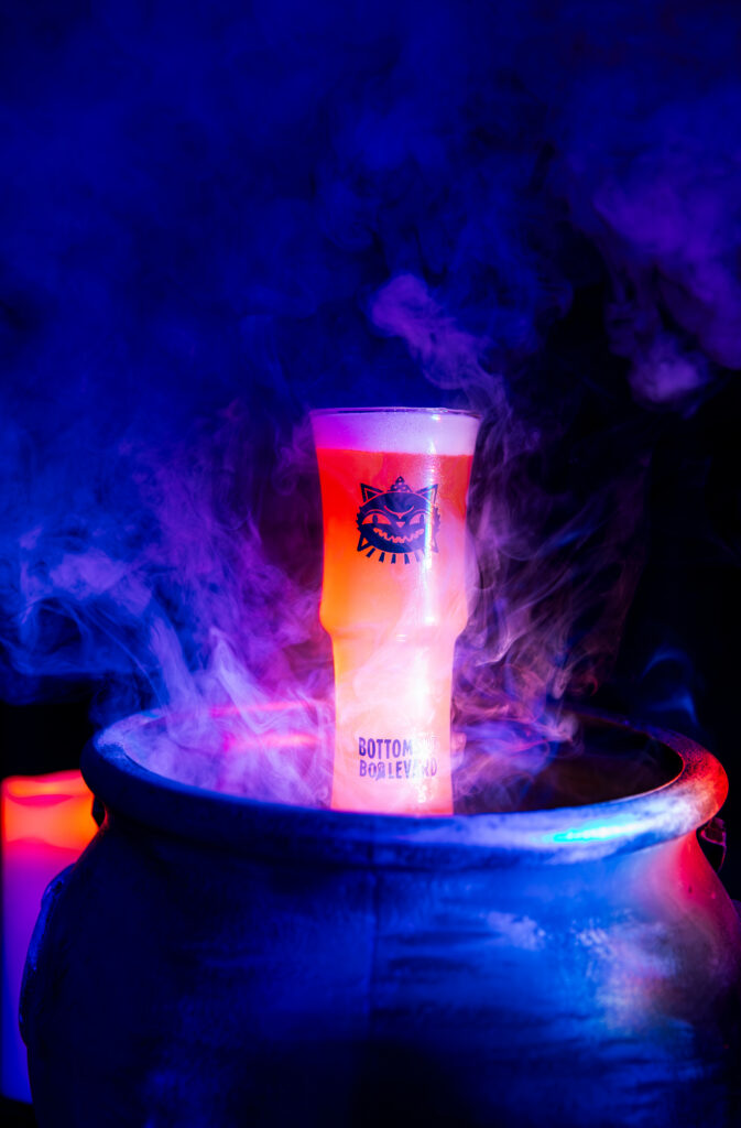 A tall, orange Boolevard cocktail in a glass with a cat logo sits inside a dark cauldron surrounded by blue and purple smoke.