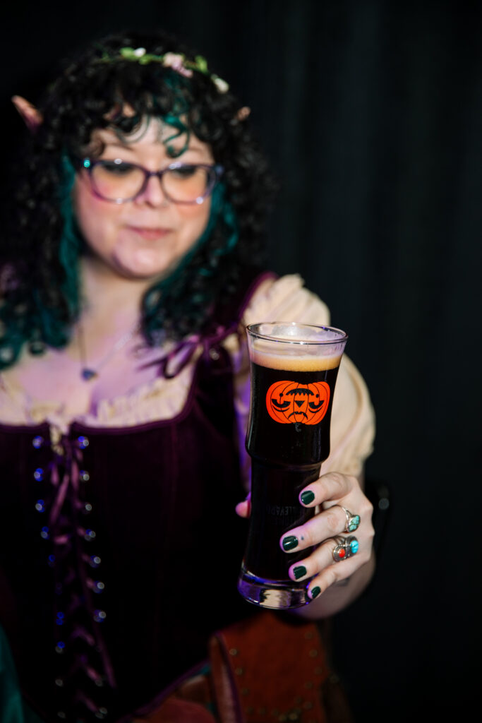 A person in costume holds up a Boulevard glass of dark beer featuring an orange jack-o’-lantern design.
