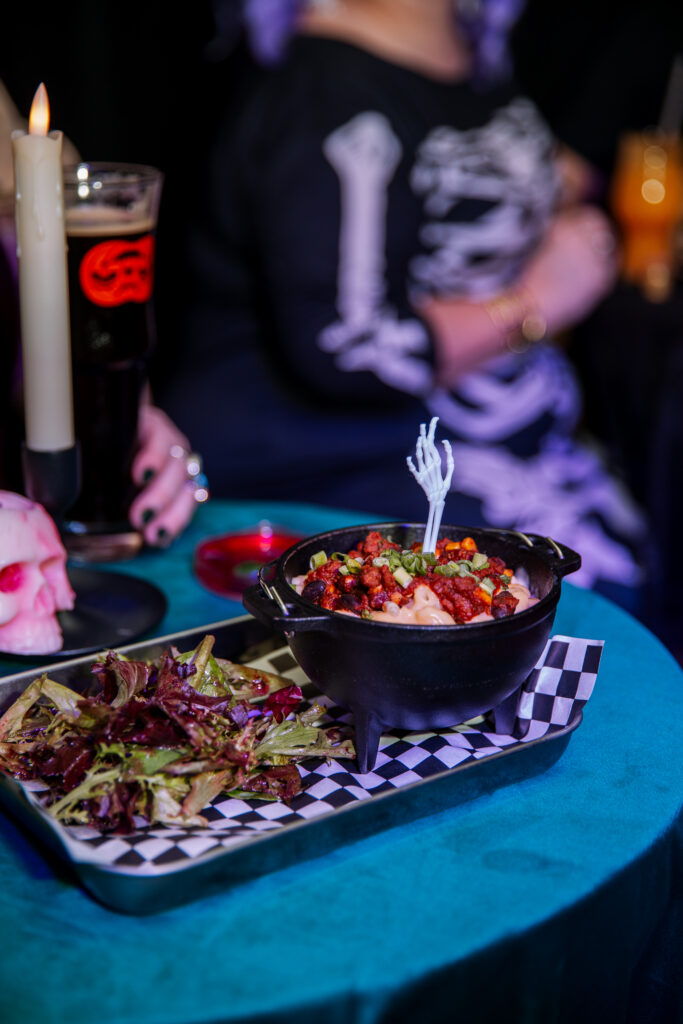 A cast iron cauldron of mac and cheese topped with crumbled sausage, garnished with a skeleton hand pick and served with mixed greens on a Boolevard checkerboard paper tray.