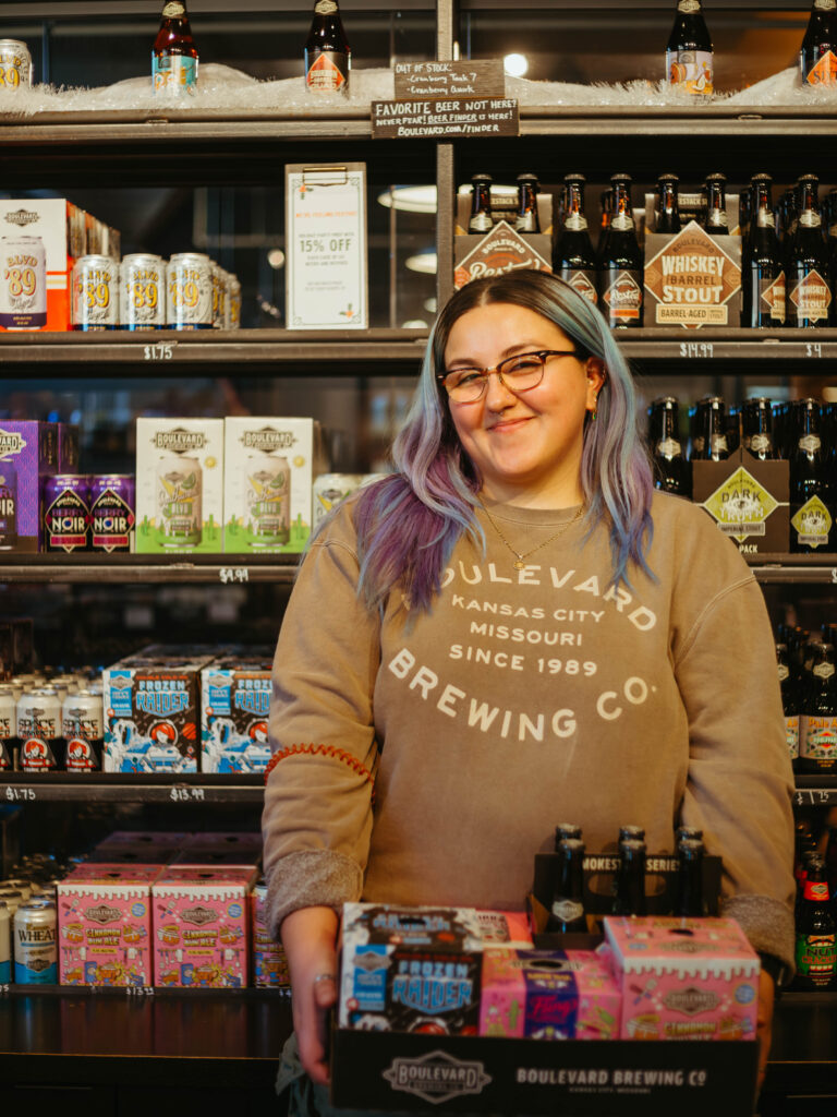 A smiling person with blue and purple hair wearing a Boulevard Brewing Company sweatshirt holds a box of beer in a store aisle lined with shelves of craft beer.