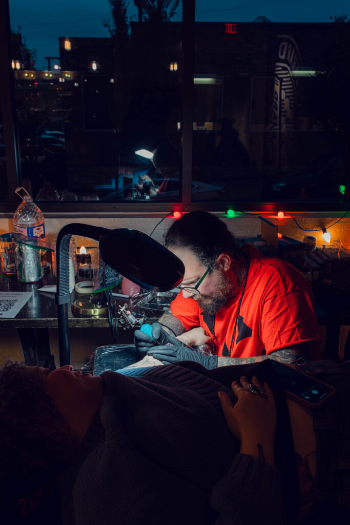 A tattoo artist in an orange shirt works on a client’s arm in a dim, Freaky Friday-inspired studio.