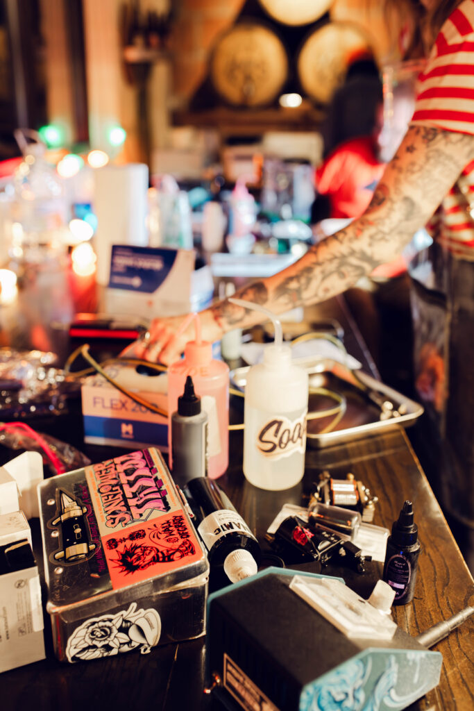 A tattoo artist with tattooed arms prepares equipment, including ink bottles, soap, and Freaky Friday tattoo machines on a table.